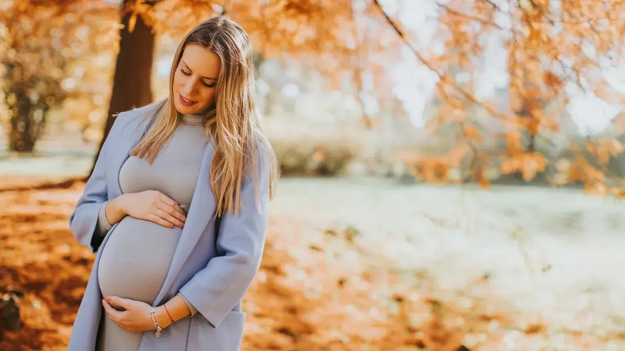 Ciążowe stylizacje na zimę: przyszła mama w pastelowym płaszczu i sukience czule obejmuje brzuszek na tle jesiennego parku.