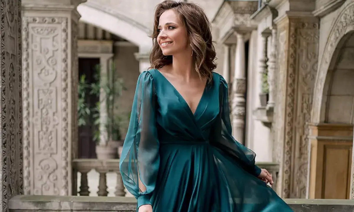 Kobieta w eleganckiej, turkusowej sukni, idealnej na coctail attire, uśmiecha się na tle zabytkowej architektury.