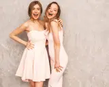 Dwie uśmiechnięte kobiety w różowych sukienkach, idealnych na cocktail attire, pozują na tle szarej ściany.