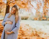Ciążowe stylizacje na zimę: przyszła mama w pastelowym płaszczu i sukience czule obejmuje brzuszek na tle jesiennego parku.
