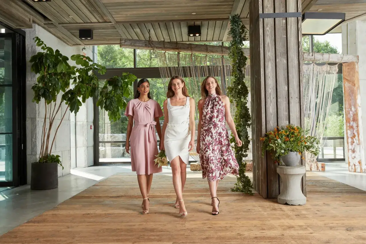 Trzy kobiety w eleganckich sukienkach prezentują styl coctail attire. Jedna w pudrowej, druga w białej, trzecia w kwiecistej kreacji.