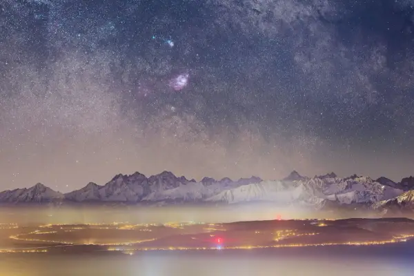 Gwieździste niebo uchwycone obiektywem do astrofotografii nad ośnieżonymi górami i rozświetlonym miastem.