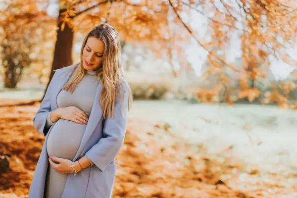 Ciążowe stylizacje na zimę: przyszła mama w pastelowym płaszczu i sukience czule obejmuje brzuszek na tle jesiennego parku.