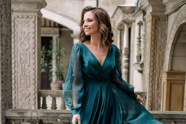 Kobieta w eleganckiej, turkusowej sukni, idealnej na coctail attire, uśmiecha się na tle zabytkowej architektury.