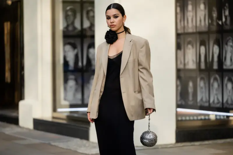 Elegancka kobieta w czarnej sukience i beżowej marynarce, z czarną różą na szyi. Idealne impreza firmowa stylizacje.